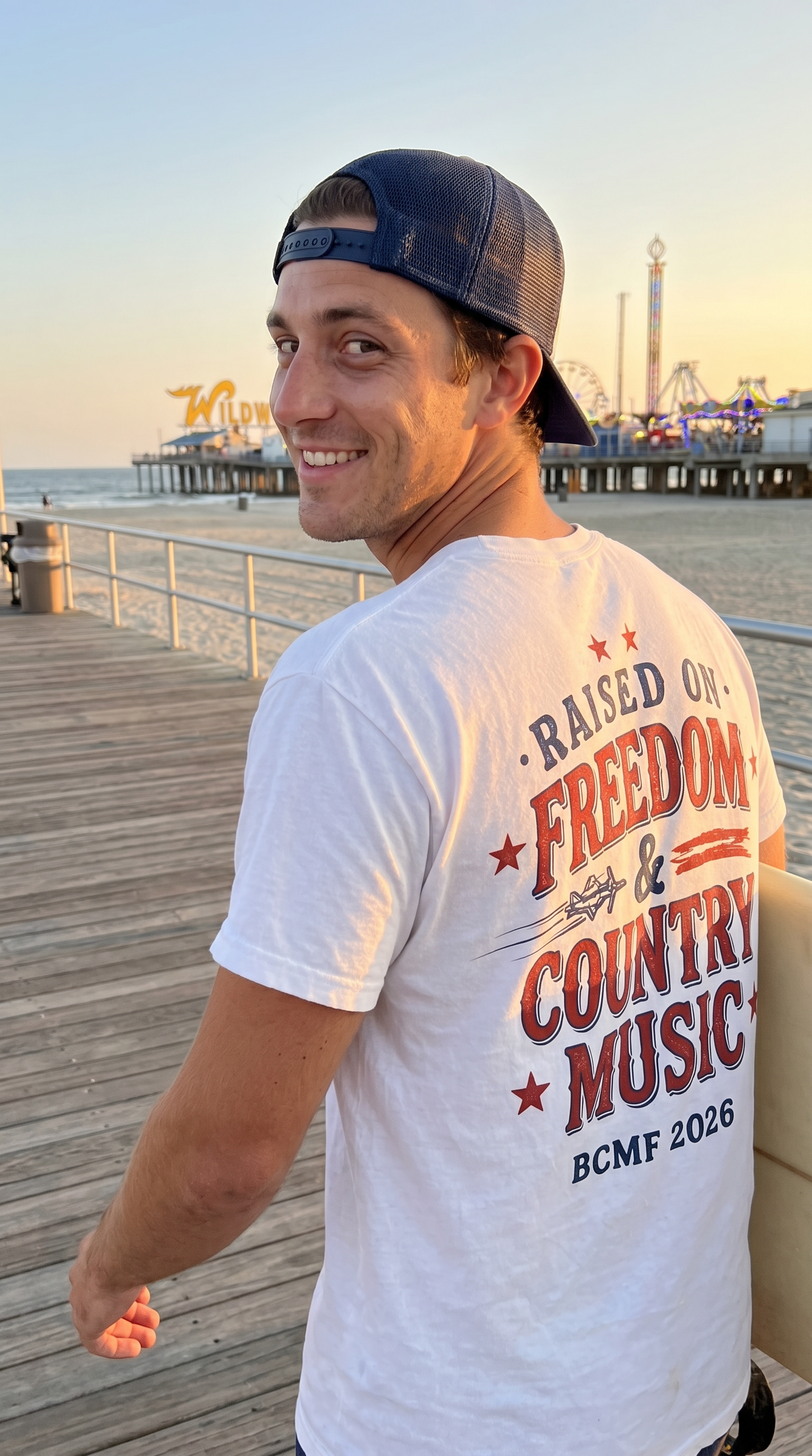 Man wearing white Raised on Freedom & Country Music back print tee on Wildwood boardwalk at golden hour